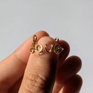 May include: A gold-coloured wire pendant spelling out the word "love" in cursive script. The pendant is held between fingers, showcasing its delicate design and warm metallic hue. The word "love" is a popular theme for jewellery.