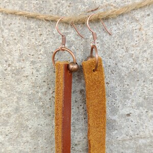 Peut inclure: Une paire de longues boucles d'oreilles en cuir marron avec des anneaux et des perles en cuivre. Les boucles d'oreilles sont suspendues à une ficelle.