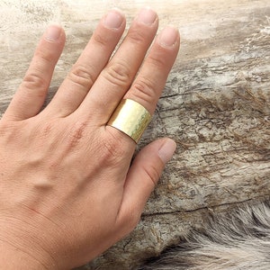 Pode incluir: Um anel largo de metal martelado dourado. O anel é usado no dedo médio de uma mão que está apoiada em um pedaço de madeira à deriva.