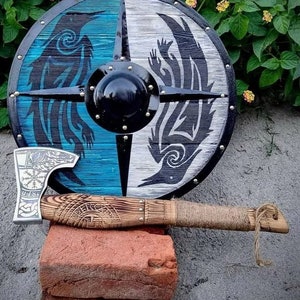 May include: A round wooden shield with a black and white design of two stylized wolves. The shield has a black center and is decorated with metal studs. A small axe with a wooden handle is resting on a brick.