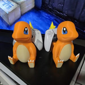 May include: Two orange Charmander figures with cream-colored bellies and blue eyes, each designed as a controller stand. The figures are holding a white and gray gaming controller, with a small yellow flame detail visible behind each controller.