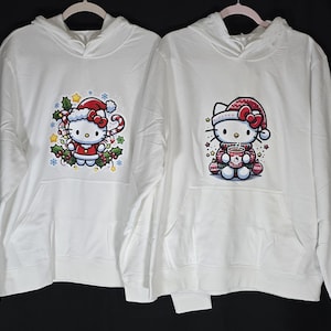Może przedstawiać: Dwie białe bluzy z kapturem z świątecznym wzorem Hello Kitty. Jedna bluza przedstawia Hello Kitty trzymającą laskę cukrową, a druga Hello Kitty trzymającą kubek gorącej czekolady.