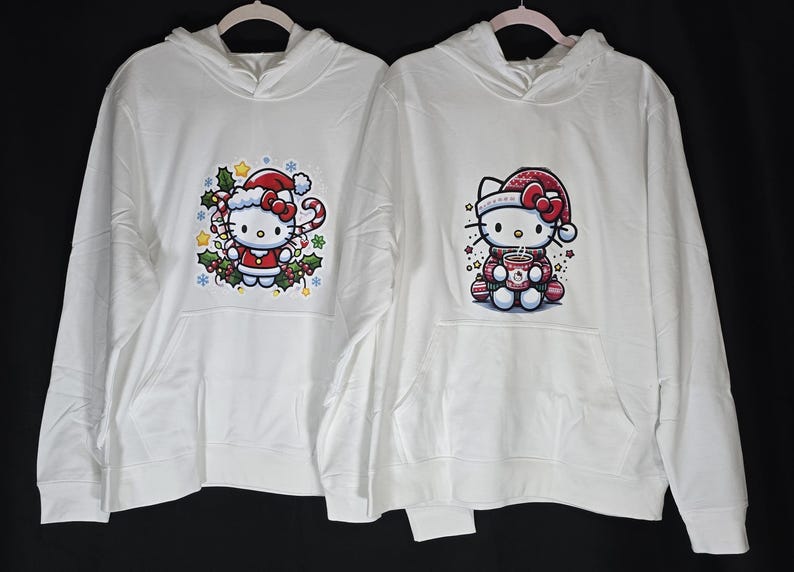 Może przedstawiać: Dwie białe bluzy z kapturem z świątecznym wzorem Hello Kitty. Jedna bluza przedstawia Hello Kitty trzymającą laskę cukrową, a druga Hello Kitty trzymającą kubek gorącej czekolady.