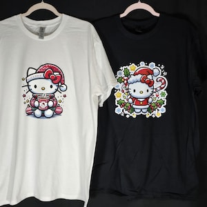 Peut inclure: Deux t-shirts avec une image de dessin anim&eacute; de Hello Kitty v&ecirc;tue d'un bonnet et d'un pull de No&euml;l rouge et blanc. Le t-shirt de gauche est blanc avec Hello Kitty tenant une tasse de chocolat chaud. Le t-shirt de droite est noir avec Hello Kitty debout dans une couronne de houx et de baies.