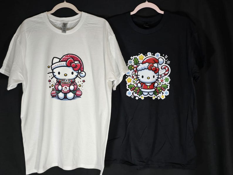 Peut inclure: Deux t-shirts avec une image de dessin anim&eacute; de Hello Kitty v&ecirc;tue d'un bonnet et d'un pull de No&euml;l rouge et blanc. Le t-shirt de gauche est blanc avec Hello Kitty tenant une tasse de chocolat chaud. Le t-shirt de droite est noir avec Hello Kitty debout dans une couronne de houx et de baies.