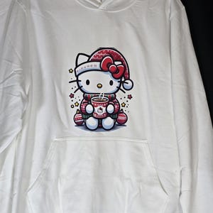 Może przedstawiać: Biała bluza z kapturem z grafiką Hello Kitty w świątecznym stylu. Hello Kitty ma na sobie czerwoną czapkę Mikołaja i trzyma czerwoną i białą filiżankę. Grafika jest otoczona małymi czerwonymi i białymi ozdobami oraz ż&oacute;łtymi gwiazdami.