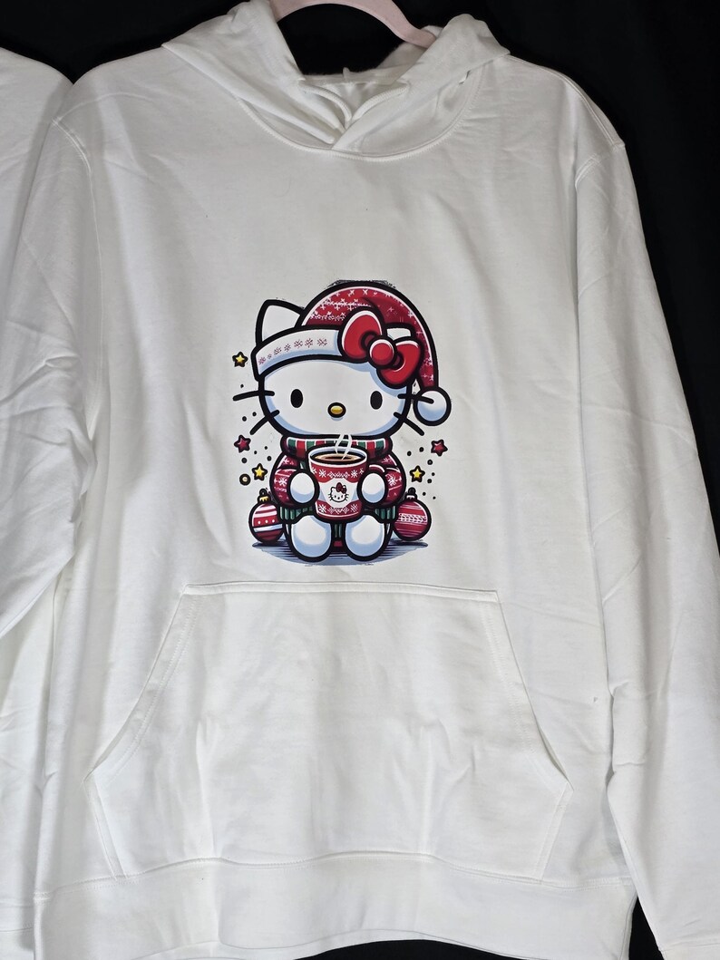 Może przedstawiać: Biała bluza z kapturem z grafiką Hello Kitty w świątecznym stylu. Hello Kitty ma na sobie czerwoną czapkę Mikołaja i trzyma czerwoną i białą filiżankę. Grafika jest otoczona małymi czerwonymi i białymi ozdobami oraz ż&oacute;łtymi gwiazdami.