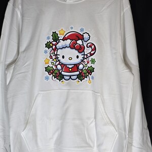 Może przedstawiać: Biała bluza z kapturem z grafiką Hello Kitty w świątecznym stylu. Hello Kitty ma na sobie czapkę Świętego Mikołaja i trzyma laskę cukrową. Grafika jest otoczona ostrokrzewem, śnieżynkami i gwiazdami.