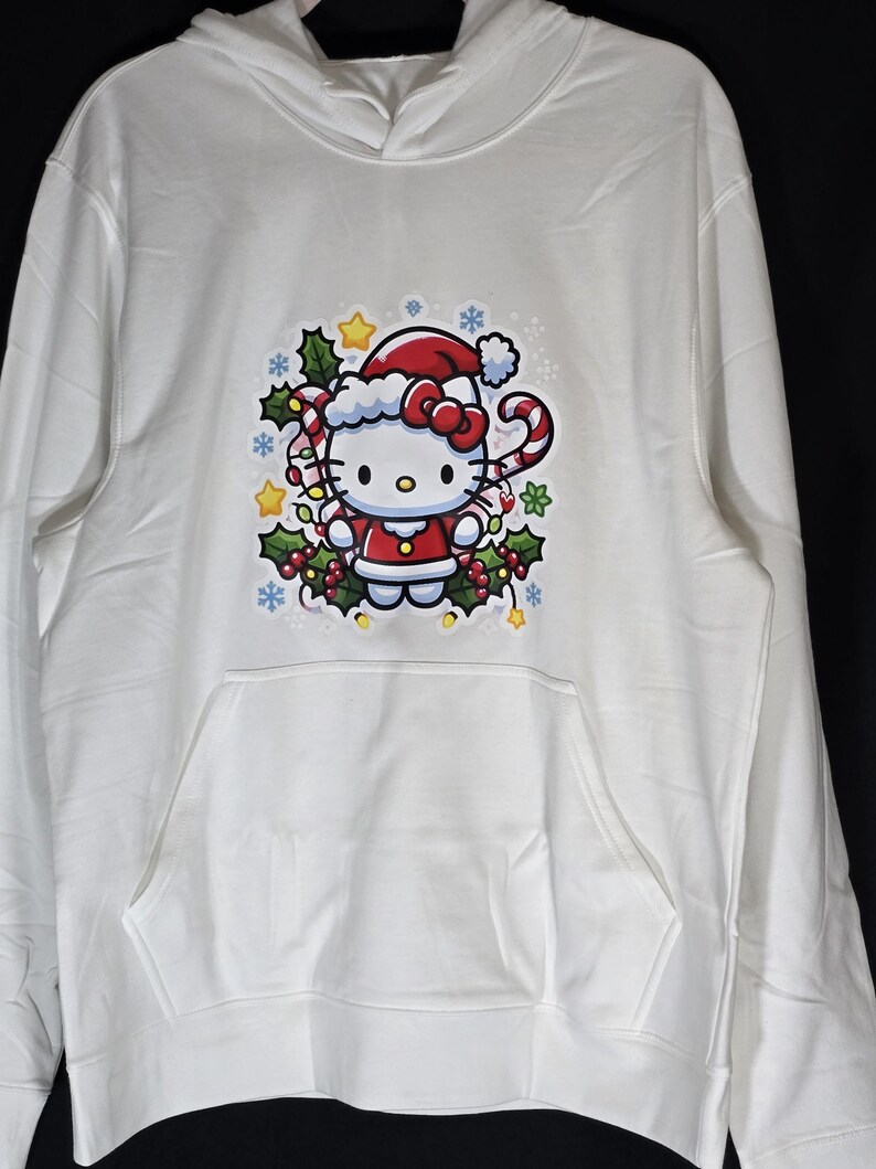 Może przedstawiać: Biała bluza z kapturem z grafiką Hello Kitty w świątecznym stylu. Hello Kitty ma na sobie czapkę Świętego Mikołaja i trzyma laskę cukrową. Grafika jest otoczona ostrokrzewem, śnieżynkami i gwiazdami.