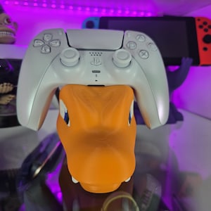 May include: A white PlayStation 5 controller resting on an orange Charmander figure. The controller has a sleek design with buttons and joysticks. A Nintendo Switch console is visible in the background, along with other gaming accessories.