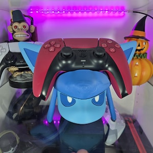May include: A red and black PlayStation 5 controller rests atop a blue figurine with large ears. A white controller and a monkey figurine are visible in the background. A Halloween pumpkin decoration is also present.