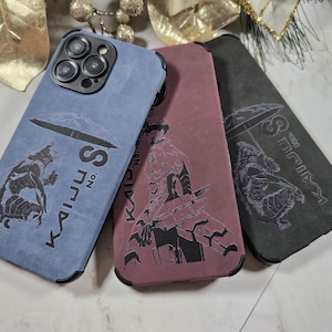 May include: Three phone cases in blue, mauve, and charcoal gray, each featuring a unique black graphic design and the text "KAIJU No. S". The cases have black borders and are made of a textured material.