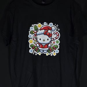 Peut inclure: T-shirt noir avec un motif blanc de Hello Kitty habill&eacute;e en P&egrave;re No&euml;l. Le motif comprend un bonnet de P&egrave;re No&euml;l rouge, une canne en sucre rouge et blanche et du houx.