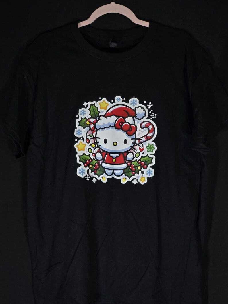Peut inclure: T-shirt noir avec un motif blanc de Hello Kitty habill&eacute;e en P&egrave;re No&euml;l. Le motif comprend un bonnet de P&egrave;re No&euml;l rouge, une canne en sucre rouge et blanche et du houx.