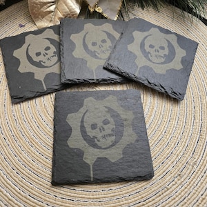 Puede incluir: Juego de cuatro posavasos cuadrados de pizarra. Cada posavasos presenta un diseño gris de calavera y engranaje. Los posavasos están colocados sobre un mantel individual beige tejido.