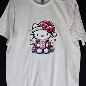 Peut inclure: T-shirt blanc avec une illustration rouge et blanche de Hello Kitty portant un bonnet de P&egrave;re No&euml;l et tenant une tasse de chocolat chaud. L'illustration est entour&eacute;e de boules rouges et dor&eacute;es et d'&eacute;toiles.