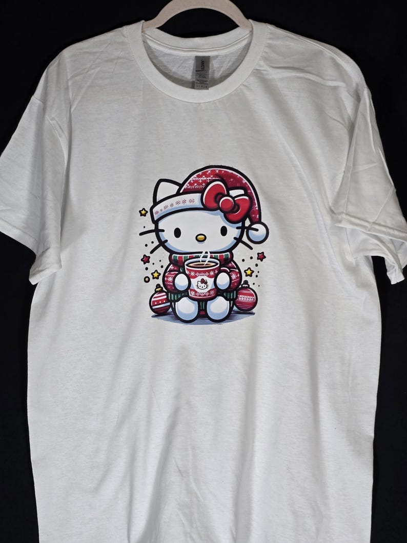 Peut inclure: T-shirt blanc avec une illustration rouge et blanche de Hello Kitty portant un bonnet de P&egrave;re No&euml;l et tenant une tasse de chocolat chaud. L'illustration est entour&eacute;e de boules rouges et dor&eacute;es et d'&eacute;toiles.