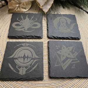 May include: Four square slate coasters with etched designs. Each coaster features a different design: a dragon, a figure with a hammer, a geometric emblem, and a floral pattern. The coasters are dark gray with a rough texture.