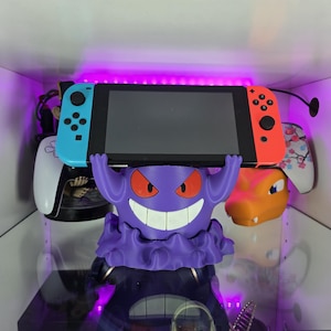 May include: A purple Gengar-themed Nintendo Switch holder with a Switch console. The Switch has blue and red Joy-Cons. Other gaming accessories are visible in the background.