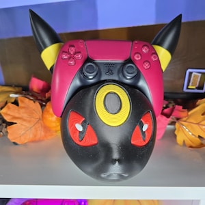 May include: A custom-designed gaming controller accessory in the shape of a black Umbreon character. The controller has a pink top with black horns and yellow accents. The Umbreon's face features red eyes and a yellow ring.