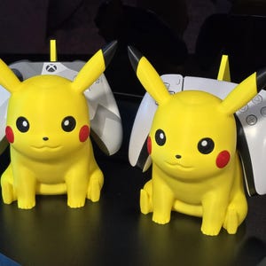 May include: Two yellow Pikachu figures with black-tipped ears and red cheek circles. Each figure is positioned in front of a white and gray game controller. The controllers have a yellow stand.