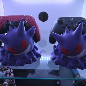 May include: Two purple and black video game controller stands shaped like the Pokemon, Gengar. The stands are holding a pink and black Playstation 5 controller and a black Xbox controller.