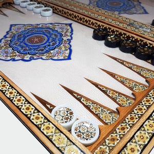 Puede incluir: Un tablero de backgammon con una superficie de juego blanca y diseños intrincados de madera incrustada. El tablero tiene un diseño floral azul y dorado en el centro. Hay fichas negras y blancas en el tablero.