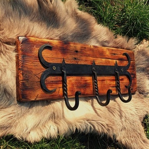 Może przedstawiać: Rusztyczny drewniany wieszak na ścianę z trzema czarnymi metalowymi haczykami. Haczyki są skręcone i mają ozdobny wzór. Drewno jest zabarwione na ciemnobrązowy kolor.