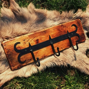 Może przedstawiać: Rusztyczny drewniany wieszak na ubrania z trzema czarnymi metalowymi haczykami. Haczyki mają kształt stylizowanej litery 'S' i mają ostry czubek. Drewno jest zabarwione na ciemnobrązowy kolor i ma naturalny rysunek drewna.