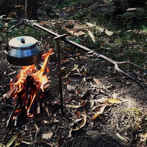 Könnte beinhalten: Ein Lagerfeuer-Setup mit einem Metallkessel, der über einem brennenden Feuer hängt. Der Kessel hängt an einem schmiedeeisernen Dreibein und Arm. Das Feuer besteht aus brennendem Holz. Die Szene spielt sich im Freien ab, mit einem natürlichen Hintergrund aus Blättern und Bäumen.