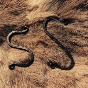Może przedstawiać: Dwa czarne metalowe haczyki w kształcie litery S z zakręconym wzorem. Haczyki leżą na brązowej, futrzanej powierzchni.