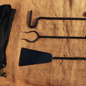 Può includere: Un set di tre strumenti per camino in metallo nero con manici intrecciati. Gli strumenti includono un attizzatoio, una pala e una pinza. Gli strumenti sono disposti su una superficie di juta marrone.