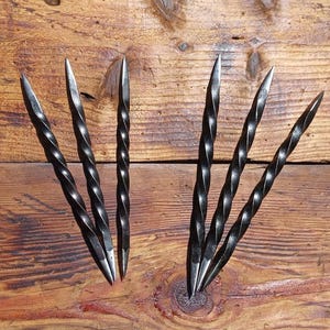 Könnte beinhalten: Sechs schwarze, handgeschmiedete Metallspieße mit einem gedrehten Design. Die Spieße haben scharfe, spitze Spitzen und sind auf einer Holzoberfläche mit einem natürlichen Maserungsmuster angeordnet. Die Spieße sind wahrscheinlich zum Grillen oder Braten gedacht.