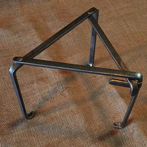 Op de afbeelding: Een driehoekige metalen standaard met drie poten. De standaard is gemaakt van stevig metaal en heeft een rustieke, industriële uitstraling.