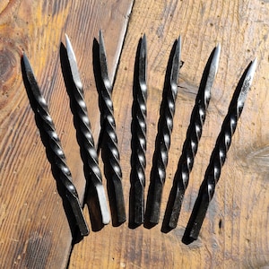 May include: A set of seven black metal skewers, each featuring a twisted, spiral-shaped shaft that tapers to a sharp, pointed tip. The metal has a dark, matte finish, and the design consists of a uniform, repeating helical pattern along the length.