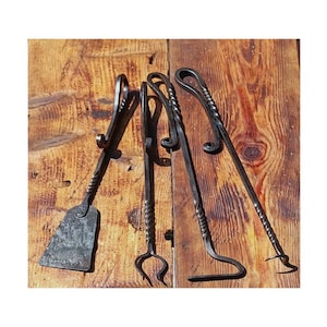 Può includere: Set di quattro attrezzi per camino artigianali in metallo nero. Include una paletta, pinze, attizzatoio e un attrezzo curvo, tutti con manici attorcigliati. Gli attrezzi sono disposti su una superficie di legno rustico.