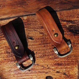 Può includere: Due portachiavi in pelle di diverse tonalità di marrone sono appoggiati su una superficie di legno. I portachiavi hanno una chiusura in metallo e una chiusura a scatto. Il legno ha un aspetto rustico e invecchiato.