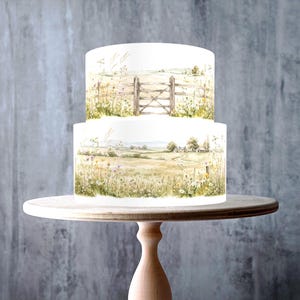 May include: A two-tier cake featuring a watercolor-style illustration of a rural landscape with a wooden gate, rolling fields, wildflowers, and a distant cottage. The design wraps around both tiers, showcasing soft green, yellow, and brown tones.