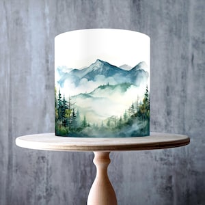 May include: A white cake with a watercolor mountain scene, displayed on a light wood cake stand. The cake features blue and green mountains with trees. The cake stand is made of light-colored wood and has a round top and a pedestal base.