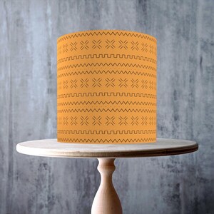 Può includere: Paralume cilindrico arancione con motivo geometrico, posto su un alzatina in legno chiaro. L'alzatina ha un piano rotondo e una base a colonna. Lo sfondo è un muro grigio strutturato. Diametro: circa 20 cm.