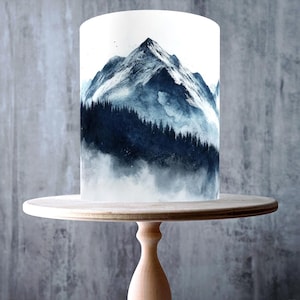 May include: A white cake with a mountain scene in shades of blue and gray, displayed on a light wood cake stand. The cake features a watercolor-style design of a mountain range and forest. The cake stand is made of light-colored wood.