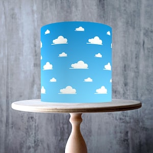 May include: A cake wrap with a blue sky and white cloud design, presented on a light wood cake stand. The stand has a circular top and a pedestal base. The image also includes a "Made in the UK" sticker and a "Vegan, Gluten Free" label.