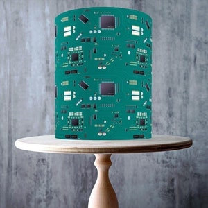 May include: A green and black circuit board pattern cake topper. The topper is on a wooden cake stand. The cake topper is made in the UK and is gluten-free, vegan, dairy-free, and nut-free.