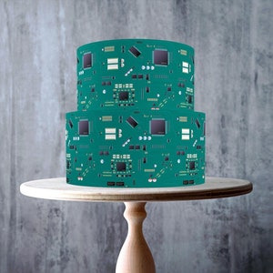 May include: A two-tiered cake decorated with a green and black circuit board design. The cake is on a wooden cake stand.
