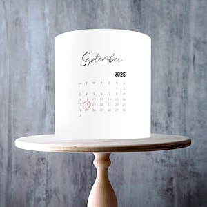 Puede incluir: Un adorno cilíndrico blanco para pasteles con un calendario impreso en negro para septiembre de 2026. La fecha 18 está resaltada con un círculo rojo. El adorno se muestra sobre un soporte de madera clara. Dos logotipos circulares en la base.
