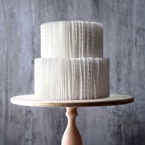 May include: A two-tiered white cake with vertical ruffled frosting texture, displayed on a light-colored wooden pedestal stand. The cake features a smooth top and sides covered in thin, pleated white icing layers, creating a uniform appearance.