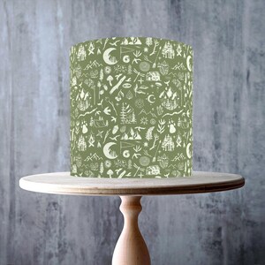 May include: A cylindrical sage green lampshade featuring a white repeating pattern of outdoor elements including pine trees, mountains, crescent moons, tents, campfires, animal tracks, and small cabins, displayed on a light wooden pedestal stand.