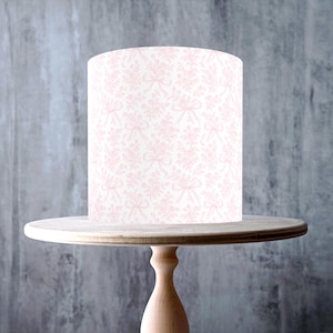 Puede incluir: Un soporte para pasteles de madera de color claro con una parte superior redonda y una base de pedestal. Un pastel cilíndrico, decorado con un patrón floral y de lazo rosa, se encuentra encima del soporte. La imagen incluye una pegatina "Made in the UK" y una pegatina "Vegan, Gluten Free, GMO Free, Nut Free, Dairy Free".