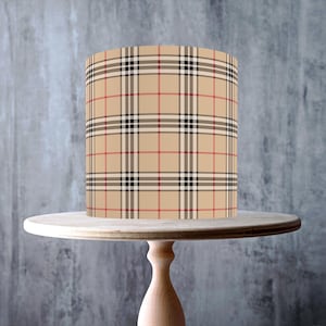 May include: A beige cake with a plaid pattern in red, black, and white, displayed on a light wood cake stand. The stand has a round top and a pedestal base. The image includes a "Made in the UK" sticker and a "Vegan" sticker.