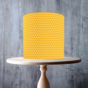 Puede incluir: Una vela cilíndrica amarilla de panal, con un patrón hexagonal repetido, se encuentra sobre un soporte para pasteles de madera clara. El soporte tiene una parte superior redonda, un pedestal torneado y una base redonda.